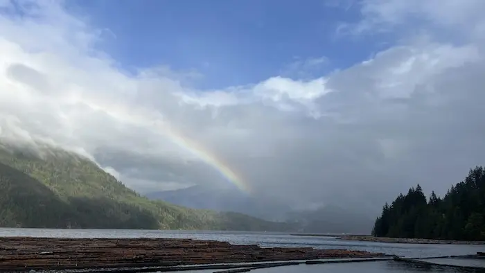Un lac avec des montagnes au fond, et en haut, un arc-en-ciel sur un fond de nuages et ciel bleu.
