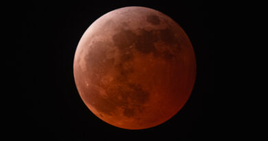 An orange-red full moon is pictured against a black night sky. The upper left of the lunar disk is relatively bright. Vast basaltic plains known as lunar maria can be seen dominating the left half of the moon's surface.