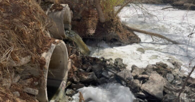 River water is shown rapidly flowing through concrete culverts, creating foam in the Tijuana River. The water appears brownish black because it is contaminated with untreated sewage.
