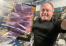 a man in a black t-shirt holds up a tray of plastic pouches filled with purple fermented milk aboard a space station.