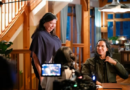 Tasha Hubbard est debout au côté de Michael Greyeyes qui est assis sur une chaise, face à une table.