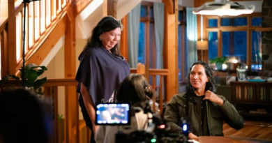 Tasha Hubbard est debout au côté de Michael Greyeyes qui est assis sur une chaise, face à une table.
