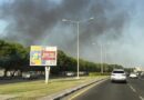 Des voitures circules sur une route. Derrière, au loin, de la fumée s'élève dans le ciel.