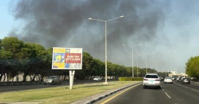 Des voitures circules sur une route. Derrière, au loin, de la fumée s'élève dans le ciel.
